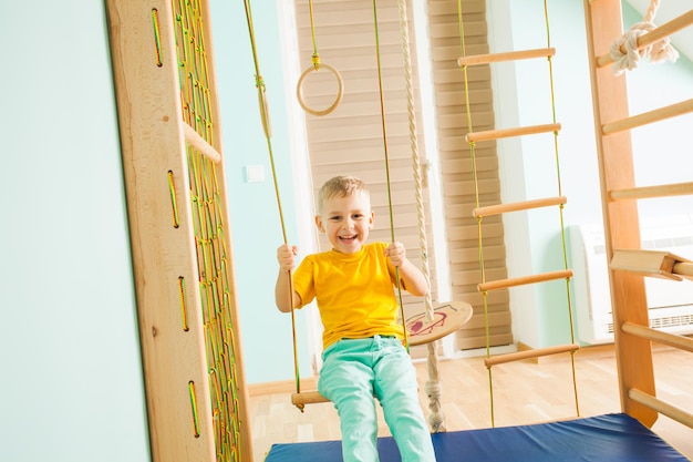 Joyful little boy sitting on a wooden swing and looking at the camera. Smiling boy in yellow t-shirt swinnging on a wooden swing at the kids home gym