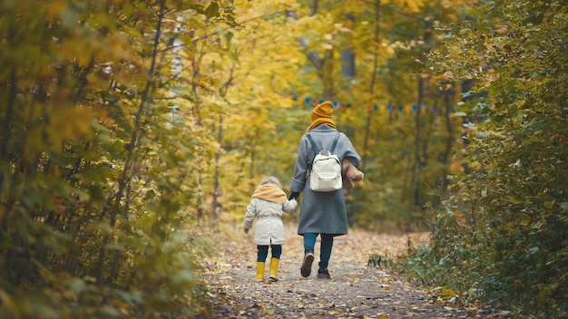 Joyful little daughter with her mommy and Teddy the Bear walks in autumn park, rear view