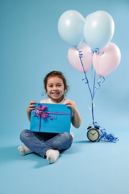 Joyful little girl cute smiling at the front sitting on a blue surface next to an alarm clock and balloons and holding a blue gift with a pink bow