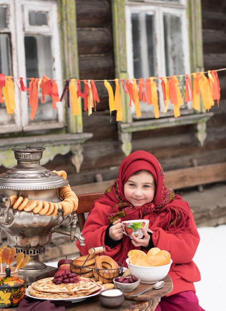 Joyful little girl drinking tea eats pankes and pies  sits at a table with a samovar