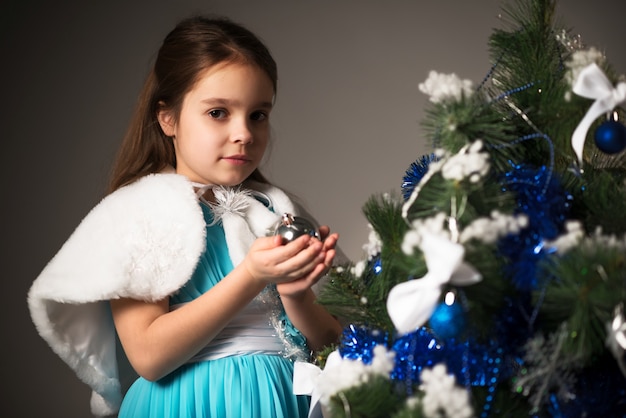 クリスマスツリーの銀のボールを保持し、灰色の背景に願い事をするうれしそうな少女。子供の頃の夢の概念と奇跡への期待。コピースペース