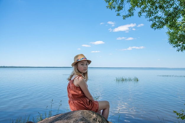 A joyful little girl in an orange dress and hat is sitting on a stone on the shore of a lake on a sunny summer day