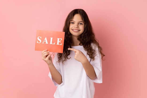 Joyful little girl pointing at card with sale inscription looking at camera with toothy smile