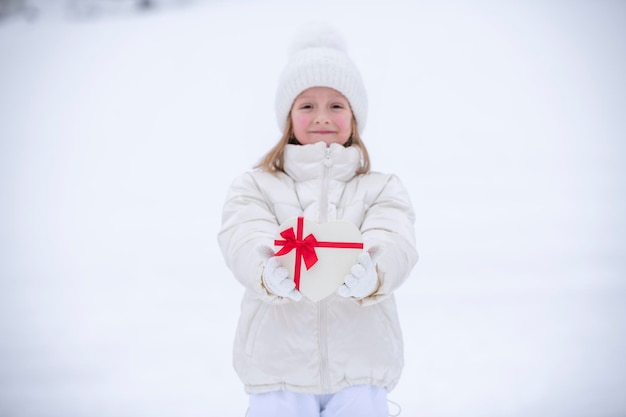 Una bambina gioiosa in abiti invernali bianchi si trova davanti alla neve e tiene in mano una scatola a forma di cuore