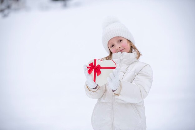 A joyful little girl in white winter clothes stands in front of the snow and holds a heartshaped box