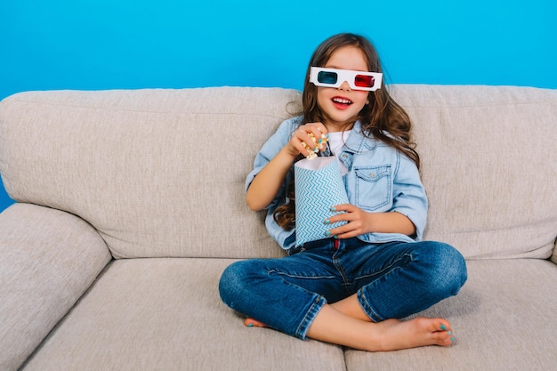 Радостная маленькая девочка с длинными волосами брюнетки в джинсовой одежде, улыбаясь в камеру с попкорном на диване, изолированном на синем фоне. Ношение 3D-очков, просмотр фильма, выражение счастья