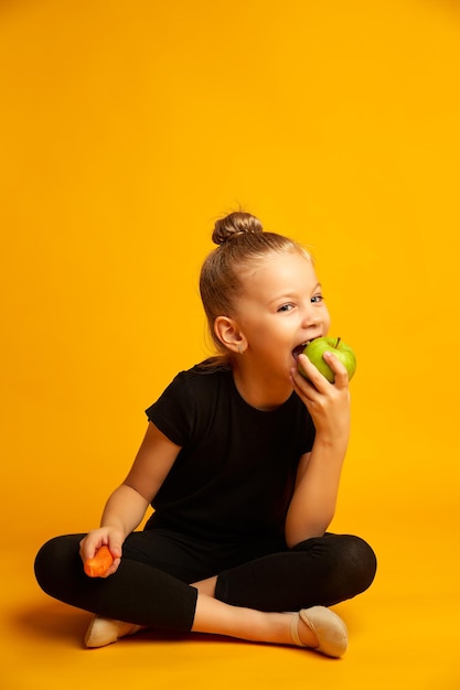 Joyful little gymnast with a carrot in his hand bites a green apple during a break studio shot on a yellow background