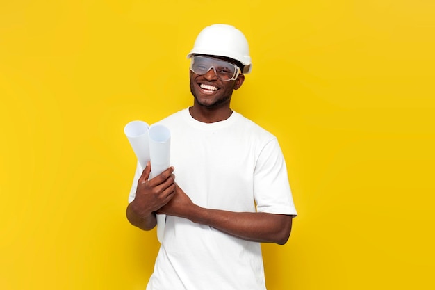 Joyful male african american builder in uniform holds drawings and projects on yellow background