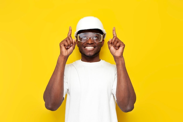 Joyful male african builder in uniform smiles and shows with his hand up on yellow background