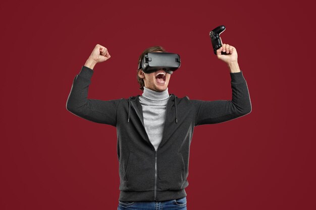 Joyful male gamer in VR headset with gamepad in hand screaming happily, and keeping fists up while rejoicing over victory in virtual game against red background