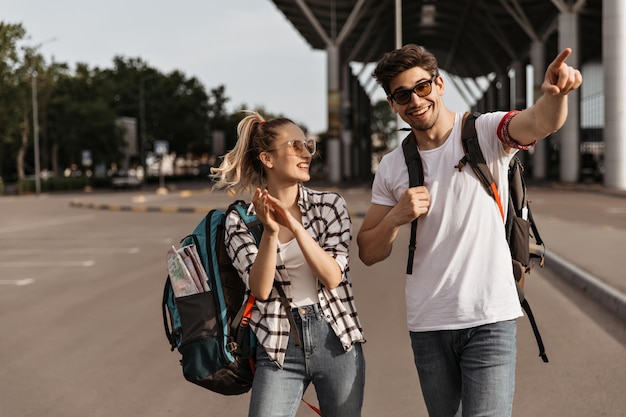 写真 ジーンズと白いtシャツのうれしそうな男と女が笑顔で話しますバックパックを持った幸せなブルネットの男と女が空港の近くを歩きます