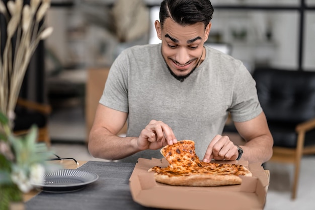 Радостный мужчина смотрит на пиццу, беря кусочек