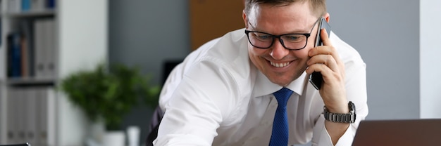 Joyful man in office speaks on phone near laptop. Introduction techniques to strengthen financial system company. Man works remotely from home during quarantine. Specialist with fresh look at work