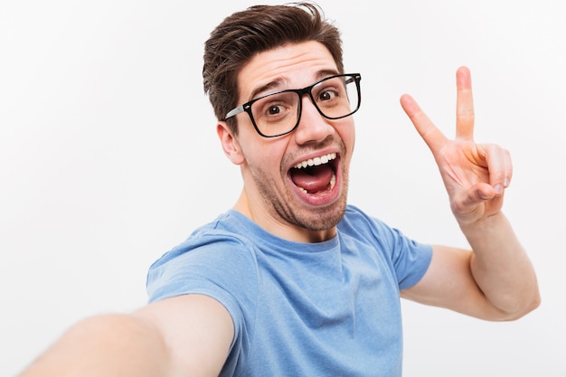 Joyful man in t-shirt and eyeglasses making selfie and showing peace gesture while looking at the camera over grey wall