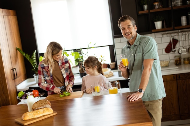 Photo joyful man with a glass of orange juice while a woman and child prepare food in a cozy kitchen