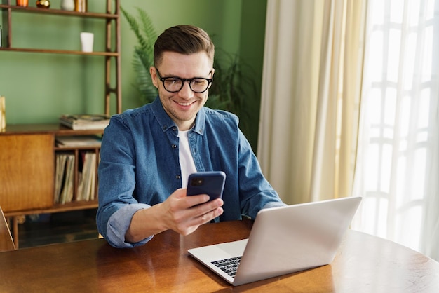 Joyful man with smartphone and laptop at sunlit worktable comfortable and stylish