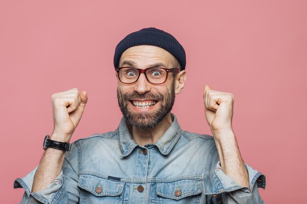 Joyful man with thick beard and mustache clenches fists and
looks joyfully at camera celebrates his victory has happy
expression isolated over pink background people emotions and
success