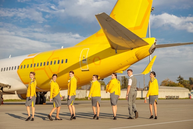 Joyful man and women flight attendants looking over shoulders and smiling while heading to airplane at airport