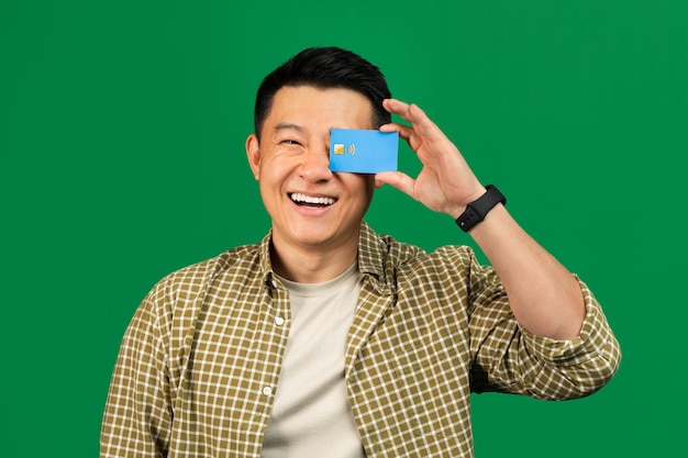 Joyful mature asian man holding credit card in front of his eye and smiling at camera male client