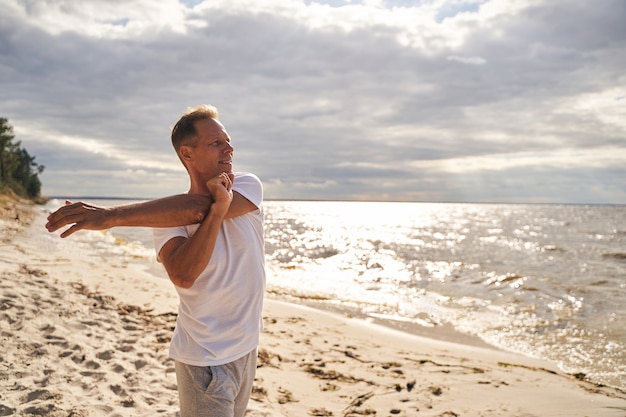 Joyful mature male is warming up before morning workout by stretching arms on sea shore