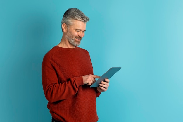 Joyful middle aged man using digital tablet on blue