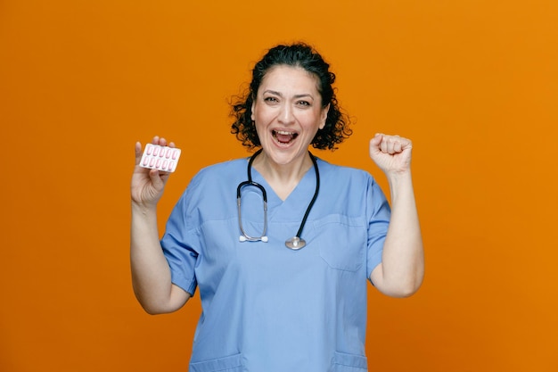 Gioiosa dottoressa di mezza età che indossa uniforme e stetoscopio intorno al collo guardando la fotocamera che mostra la confezione di capsule che mostra che lo farai gesto isolato su sfondo arancione