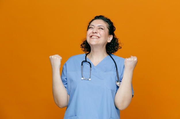 Joyful middleaged female doctor wearing uniform and stethoscope around her neck showing yes gesture with closed eyes isolated on orange background