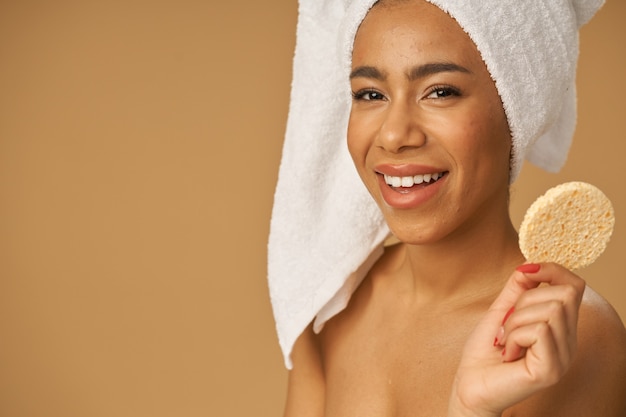 Joyful mixed race young woman holding cleansing face sponge, posing isolated over beige background