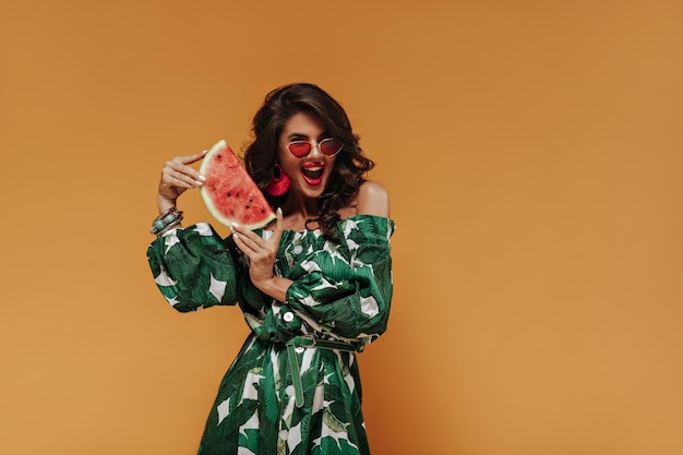 Joyful modern woman with wavy brunette hair in red earrings and white and green dress with button and thin belt having fun and holds watermelon