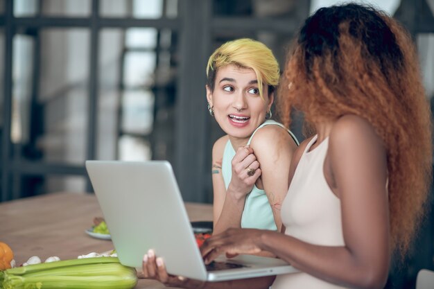 Foto momento gioioso. due giovani amiche gioiose con il computer portatile vicino al tavolo con verdure che comunicano allegra mattina in cucina