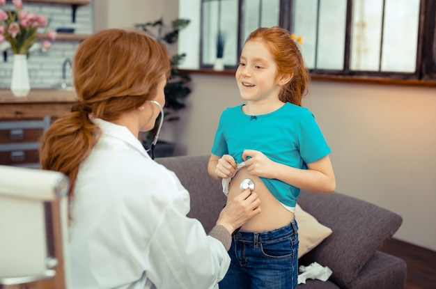 Joyful mood. Positive cute girl smiling while showing her belly to the doctor