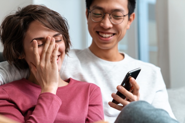 joyful multinational couple laughing and using cellphone while sitting on sofa at home