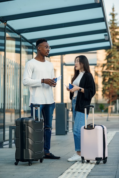 A joyful multiracial couple checks their boarding passes and departure time at a bus stop near the airport.