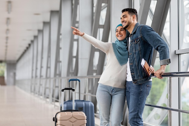 Joyful muslim couple standing with luggage at airport and pointing away