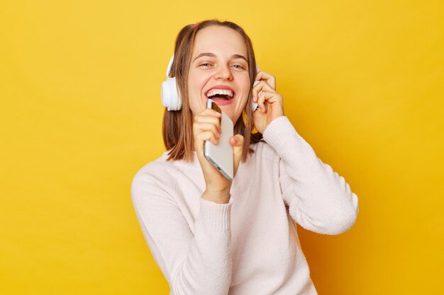 Joyful optimistic girl wearing jumper and headphones posing isolated over yellow background glad to search favorite composition on web page listening cool song in headphones singing from pleasure