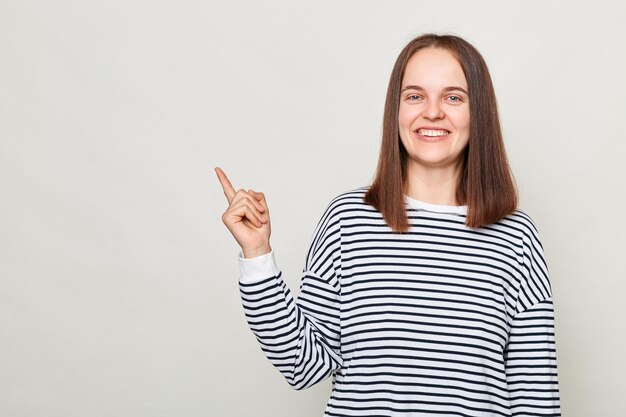 Joyful optimistic woman with brown hair wearing striped casual shirt standing isolated over gray background looking at camera showing space for your advertisement