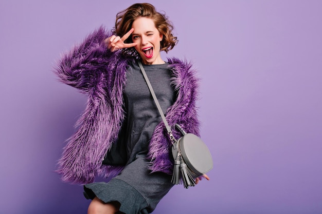 Joyful pale girl with dark curly hair dancing in purple fur coat. Studio shot of elegant pretty caucasian woman with little gray handbag.