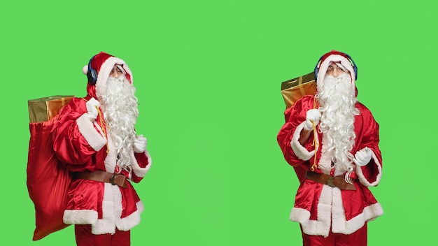 Joyful person in santa suit listen to music, having fun dancing with presents sack over greenscreen. Young adult enjoying songs on headphones, wearing festive costume and white beard.