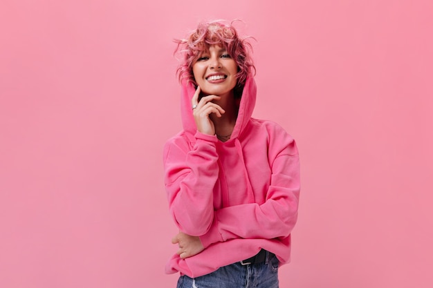 Photo joyful pink-haired curly woman in oversized hoodie and jeans smiles sincerely and touches face on isolated pink wall