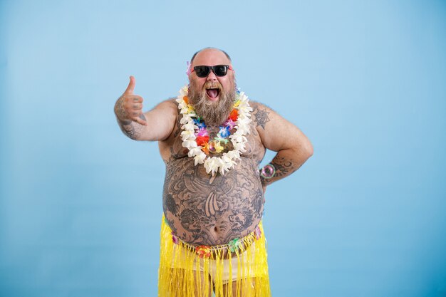 Joyful plump man with sunglasses in grass skirt shows shaka greeting gesture on blue background