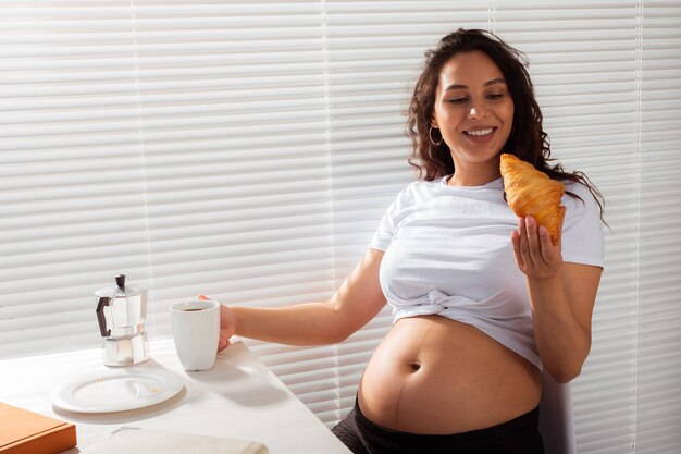 Gioiosa donna incinta mangiare croissant e bere il tè durante la colazione del mattino