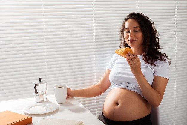 Joyful pregnant young beautiful woman eating croissant and drinking tea during morning breakfast