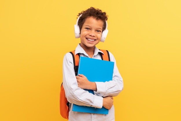 joyful preteen schoolboy with headphones and folders on yellow