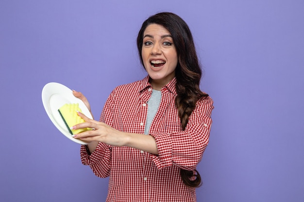 Joyful pretty caucasian cleaner woman cleaning plate with sponge and looking at front