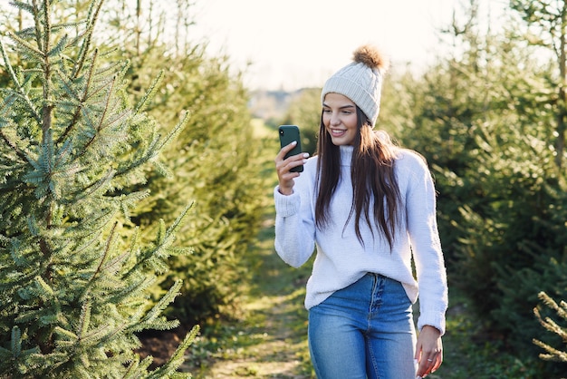 暖かい帽子の写真撮影でうれしそうなかなり若い女性は、林業植栽の間を歩いている間美しいクリスマスツリー。