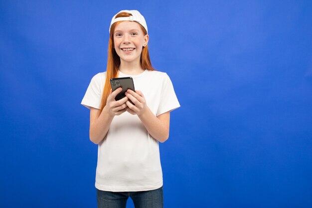 Радостная рыжеволосая девушка-подросток в белой футболке общается в социальных сетях со смартфона на синем