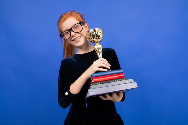 Joyful red-haired teenager girl with a stack of books and a victory cup on blue