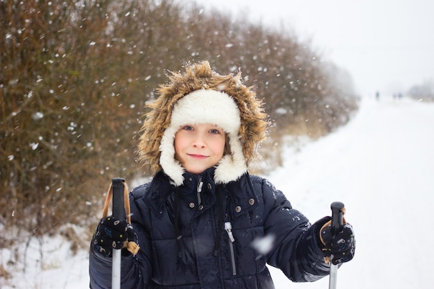 Gioioso ragazzo russo sciare in inverno durante la nevicata con bastoncini da sci sulla strada forestale.