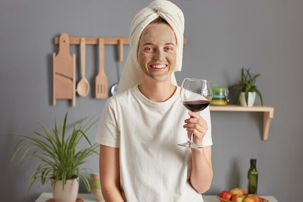 Joyful satisfied relaxed young woman posing in kitchen with towel on head drinking wine in the morning enjoying weekend smiling to camera