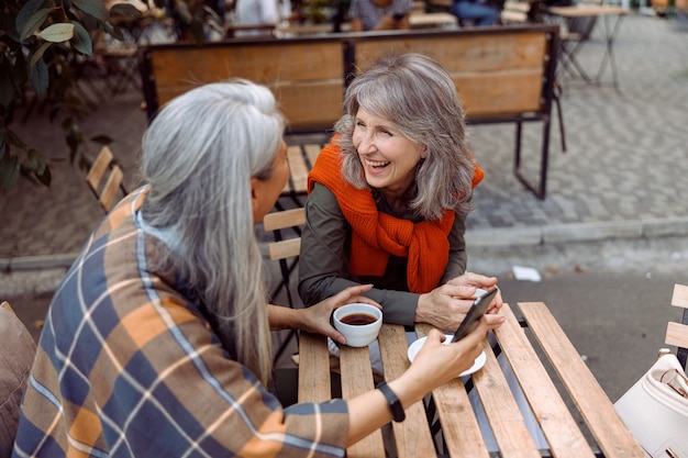 Joyful senior ladies best friends with phone and coffee sit at table in street cafe